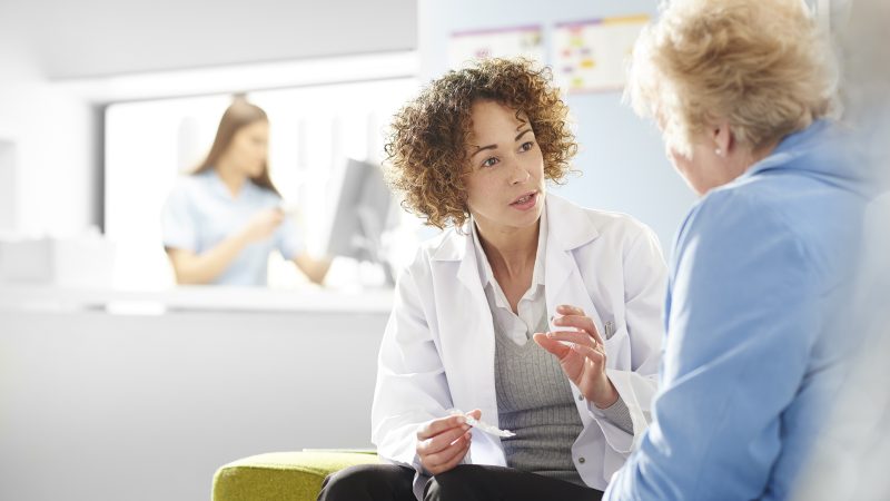 A female pharmacist sits with a senior female patient in the pharmacist consultation area and discusses her prescription and choice of medication. In the background a father and daughter stand at the dispensing counter and are served by a female pharmacy assistant .