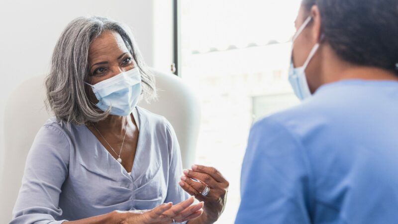 A senior woman, wearing a protective face mask, talks with a female nurse during a medical appointment.